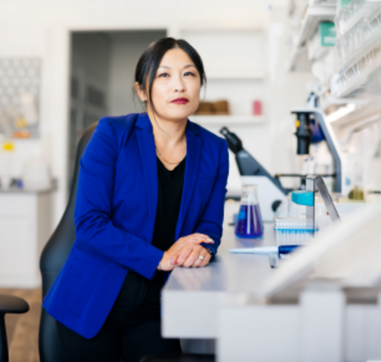 Woman of Asian descent is leaning on a laboratory counter with microscopes